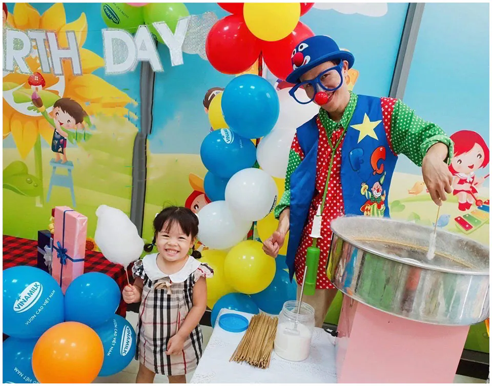 Chú hề vui nhộn đang biểu diễn làm kẹo bông gòn bằng máy chuyên dụng tặng cho bé gái dễ thương trong buổi tiệc sinh nhật có trang trí bong bóng in logo Vinamilk.