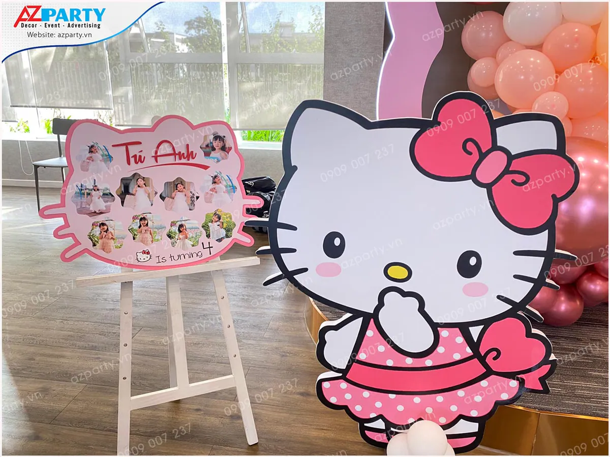 Mô hình chú mèo Hello Kitty mặc đầm hồng chấm bi dễ thương đứng cạnh khung ảnh kỷ niệm của bé Tú Anh trên giá đỡ gỗ.