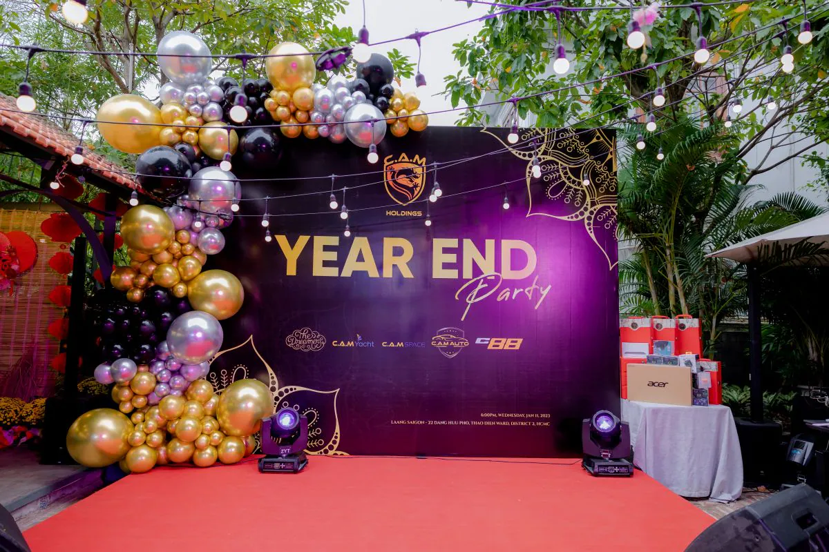Sân khấu tiệc tất niên C.A.M Holdings Year End Party với backdrop tone tím đen, trang trí bóng bay vàng bạc đen và đèn sân khấu chuyên nghiệp