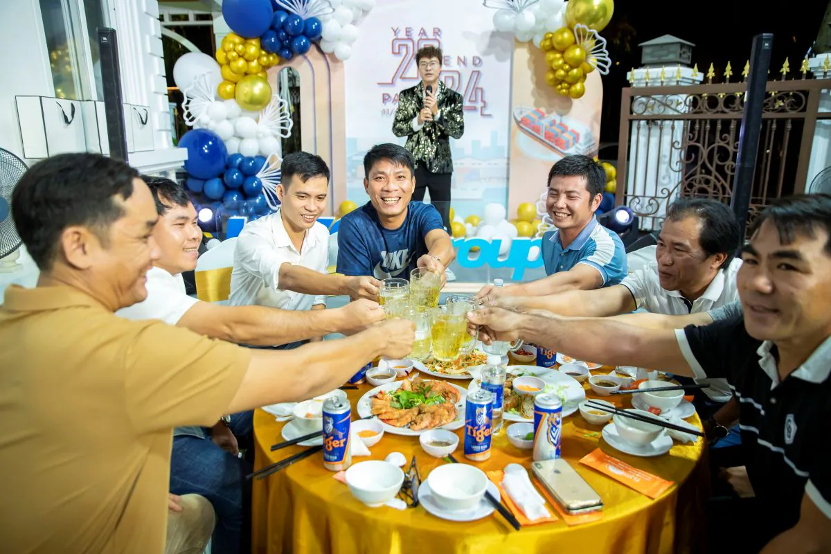 Nhóm nam nhân viên HVS Group vui vẻ cùng nâng ly chúc mừng tại bàn tiệc trong khi MC đang dẫn chương trình trên sân khấu phía sau tại sự kiện tất niên Year End Party 2024.