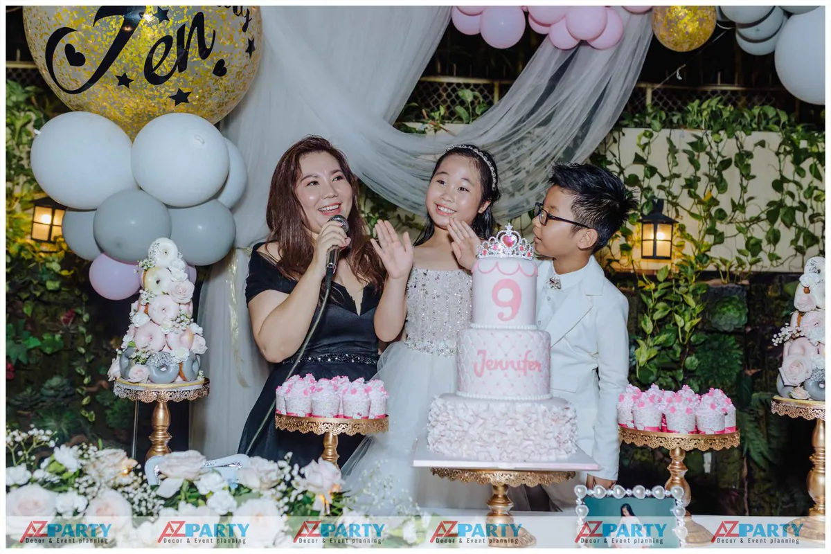 Bé Jennifer rạng rỡ đón tuổi mới bên mẹ và em trai đứng sau chiếc bánh kem sinh nhật 3 tầng chuẩn bị thổi nến chúc mừng.