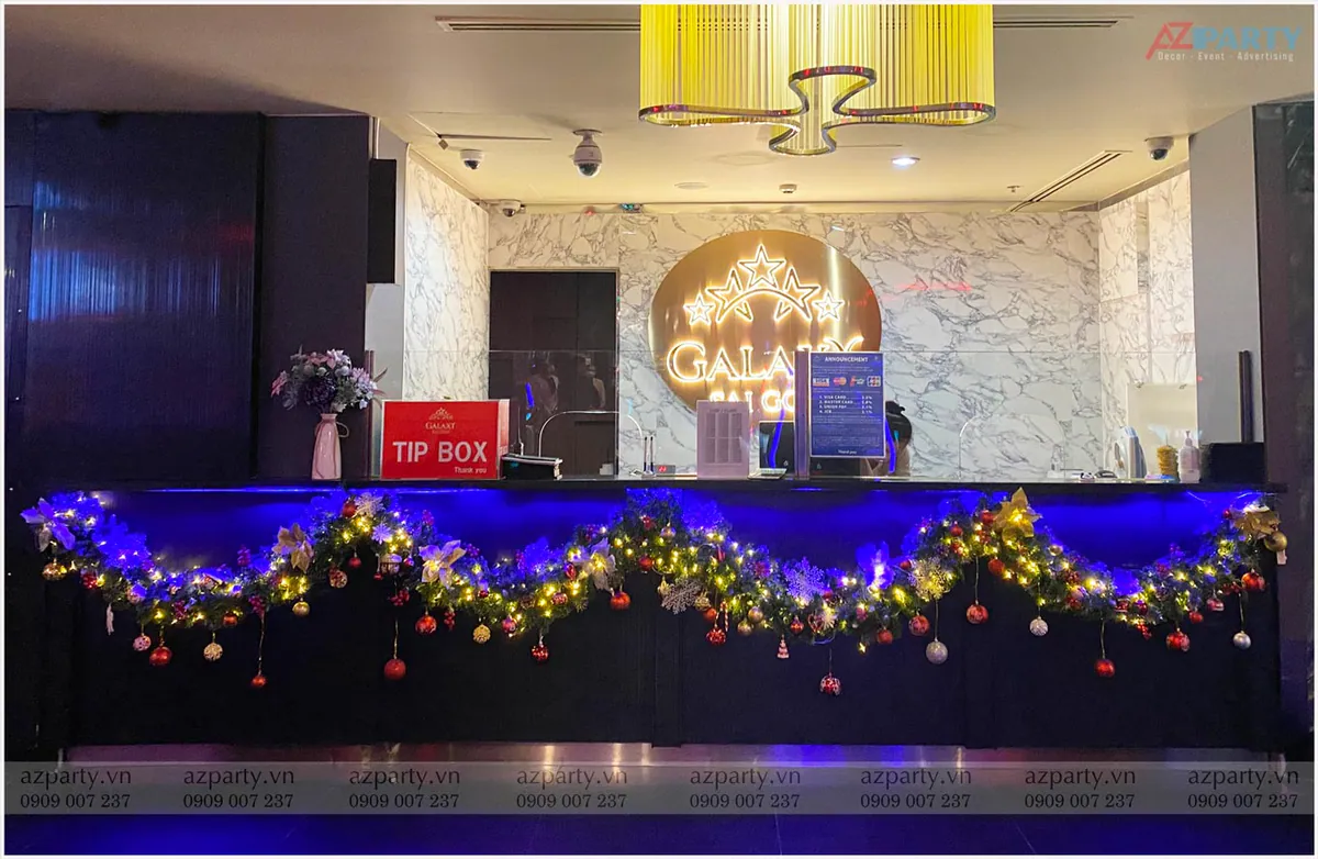 Góc trang trí Noel tại quầy lễ tân Galaxy Club với dải lá thông uốn lượn chăng đèn LED vàng ấm áp treo rủ nhiều quả châu dưới ánh đèn mờ tím ảo diệu