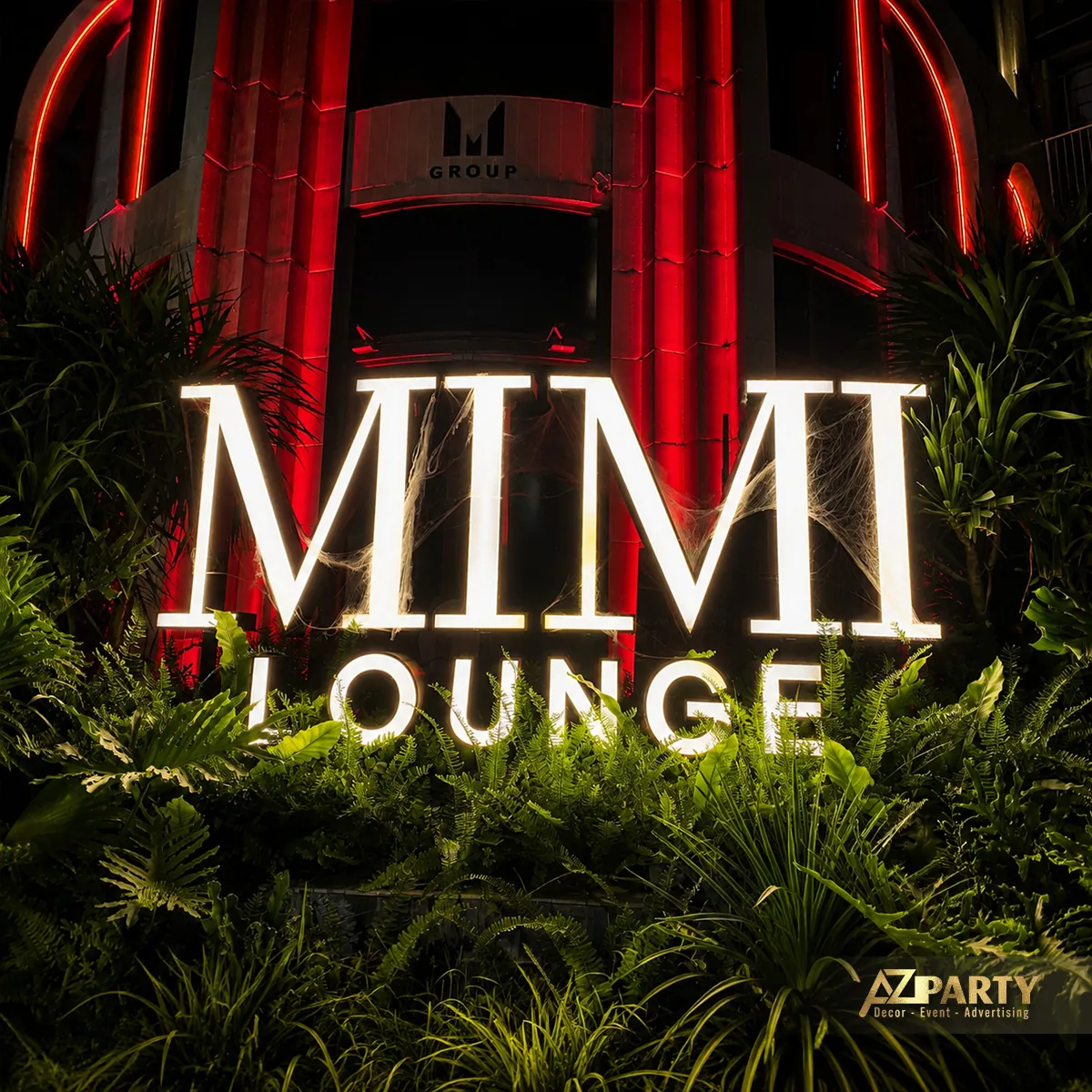 Bảng chữ MIMI LOUNGE phát sáng giữa mảng cây xanh và ánh đèn đỏ.