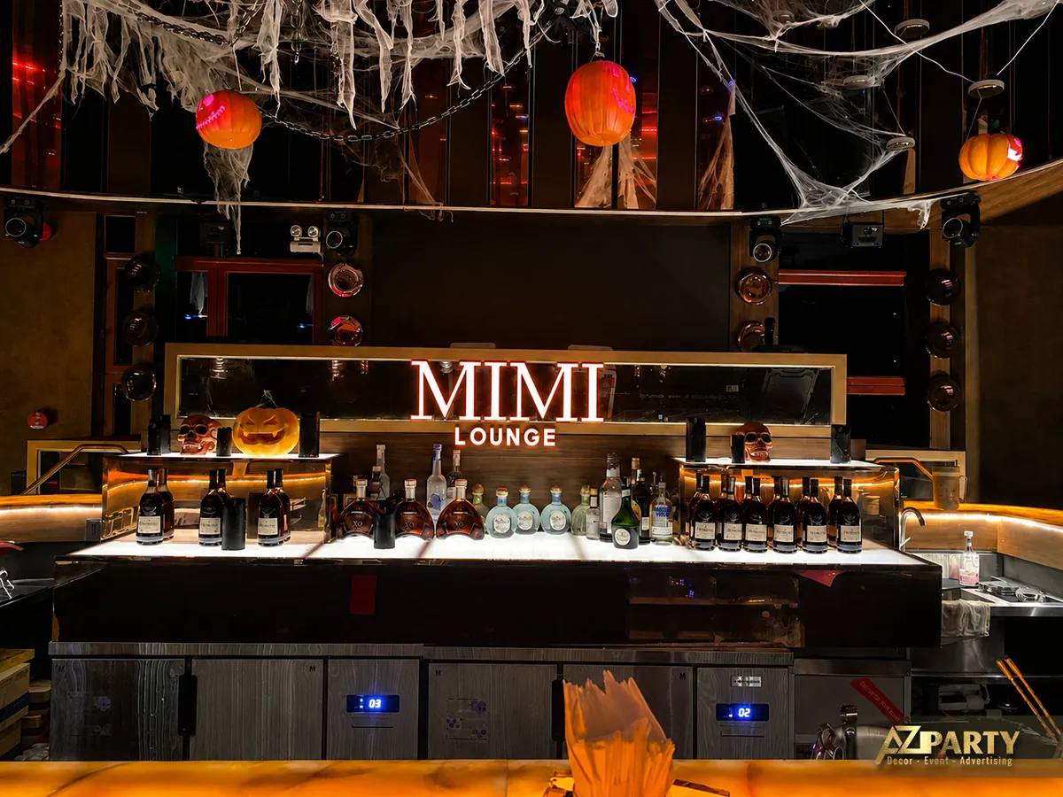 Góc chụp cận diện quầy bar Mimi Lounge với bảng tên, các chai rượu xếp trên kệ và đồ trang trí Halloween.