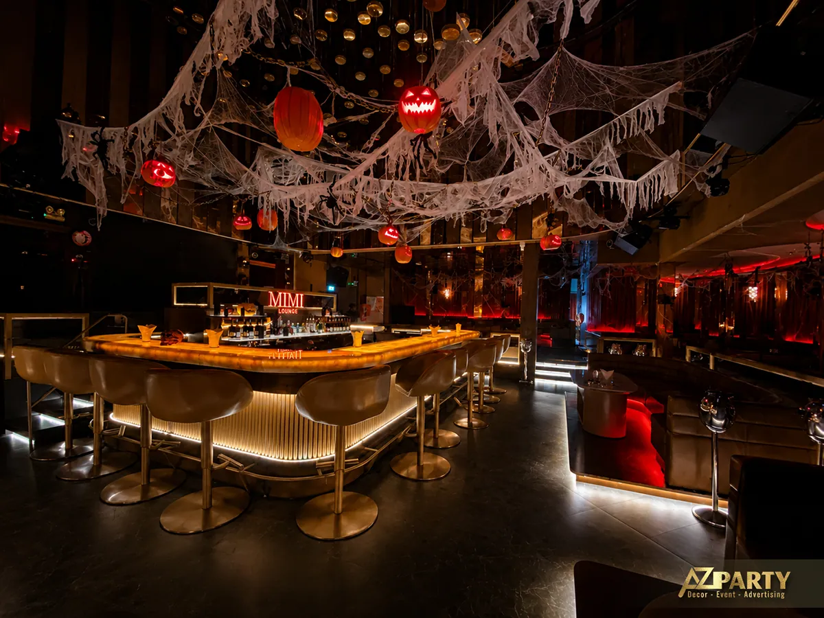Toàn cảnh quầy bar Mimi Lounge nhìn chếch từ bên phải, với ghế bar, khu vực lounge, mạng nhện và đèn bí ngô treo trên trần.