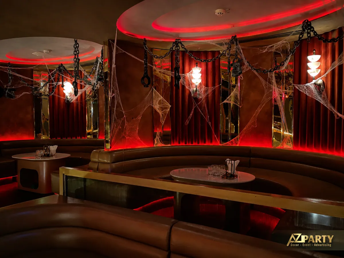 Góc ghế lounge đối diện mảng tường rèm đỏ, với mạng nhện, đèn treo và phần bàn thấp ở trung tâm.