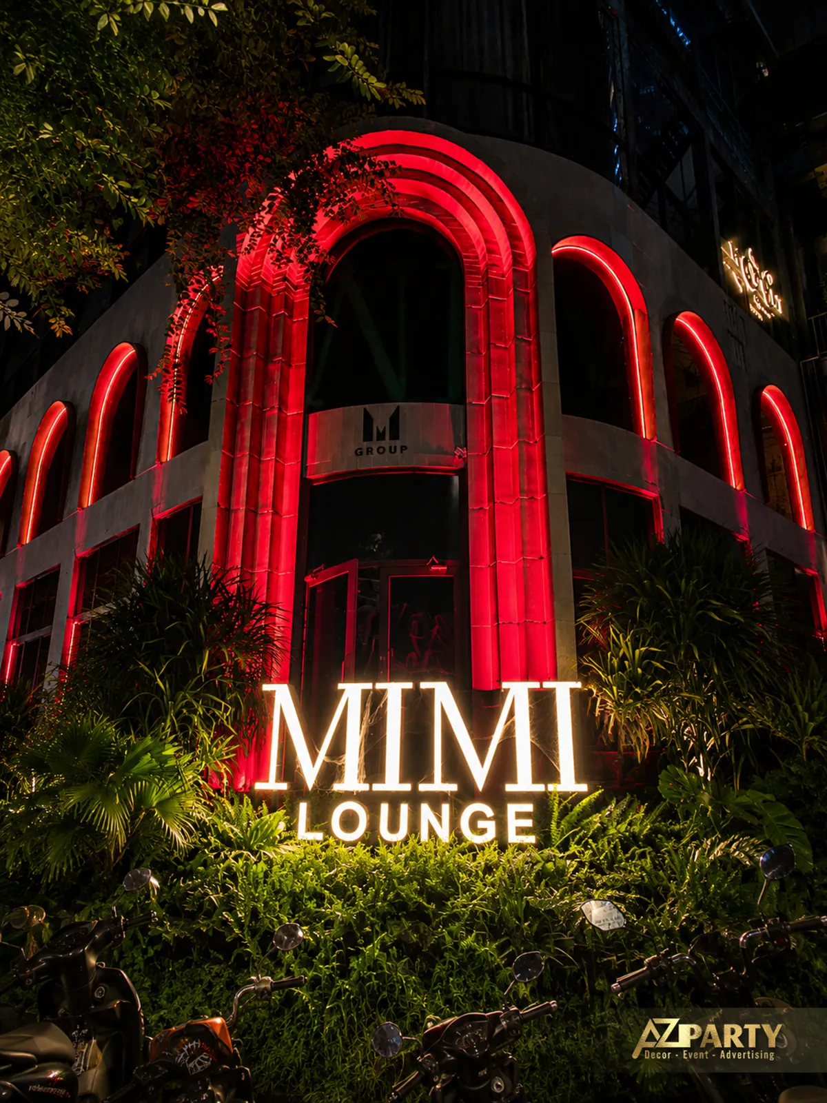 Mặt tiền Mimi Lounge với các đường viền đèn đỏ trên kiến trúc vòm, cây xanh phía trước và bảng hiệu phát sáng.