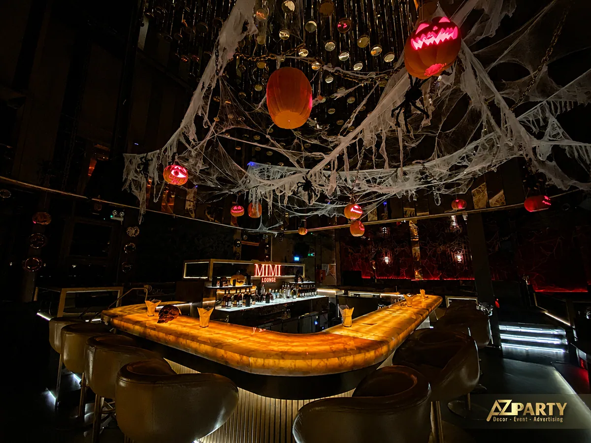 Góc chụp chếch toàn bộ quầy bar, với mặt quầy phát sáng, ghế bar và các bí ngô, mạng nhện treo phía trên.