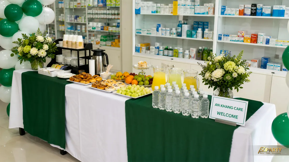 Bàn tiệc An Khang Care phủ khăn xanh trắng với bánh, trái cây, nước và hoa