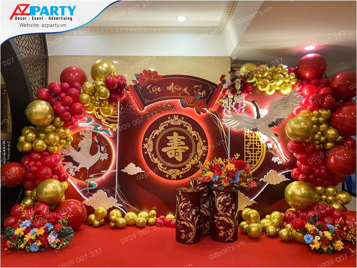 Backdrop trang trí lễ mừng thọ phong cách Trung Hoa sang trọng với tông màu đỏ, vàng gold chủ đạo, chữ 'Thọ' tiếng Hán ở trung tâm, mô hình chim hạc bay lượn và bóng bay hai bên.