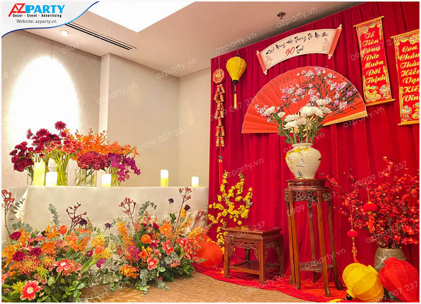Khu vực trang trí truyền thống cho lễ mừng thọ cụ bà 90 tuổi với rèm nhung đỏ, đèn lồng, quạt giấy lớn vẽ hoa đào và bình hoa cúc trắng trên đôn gỗ.
