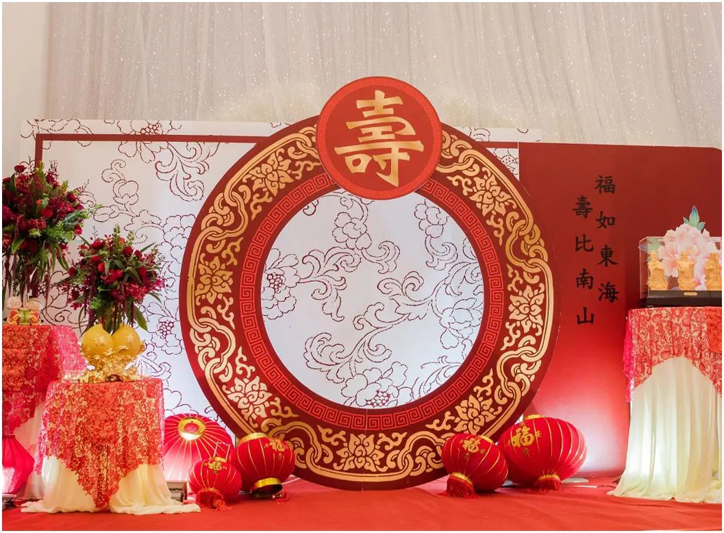 Backdrop mừng thọ với khung tròn họa tiết cổ điển Trung Hoa kết hợp câu đối 'Phúc như Đông Hải, Thọ tỷ Nam Sơn' viết bằng chữ Hán thư pháp màu đen trên nền đỏ, đi kèm đèn lồng đỏ đặt dưới sàn.