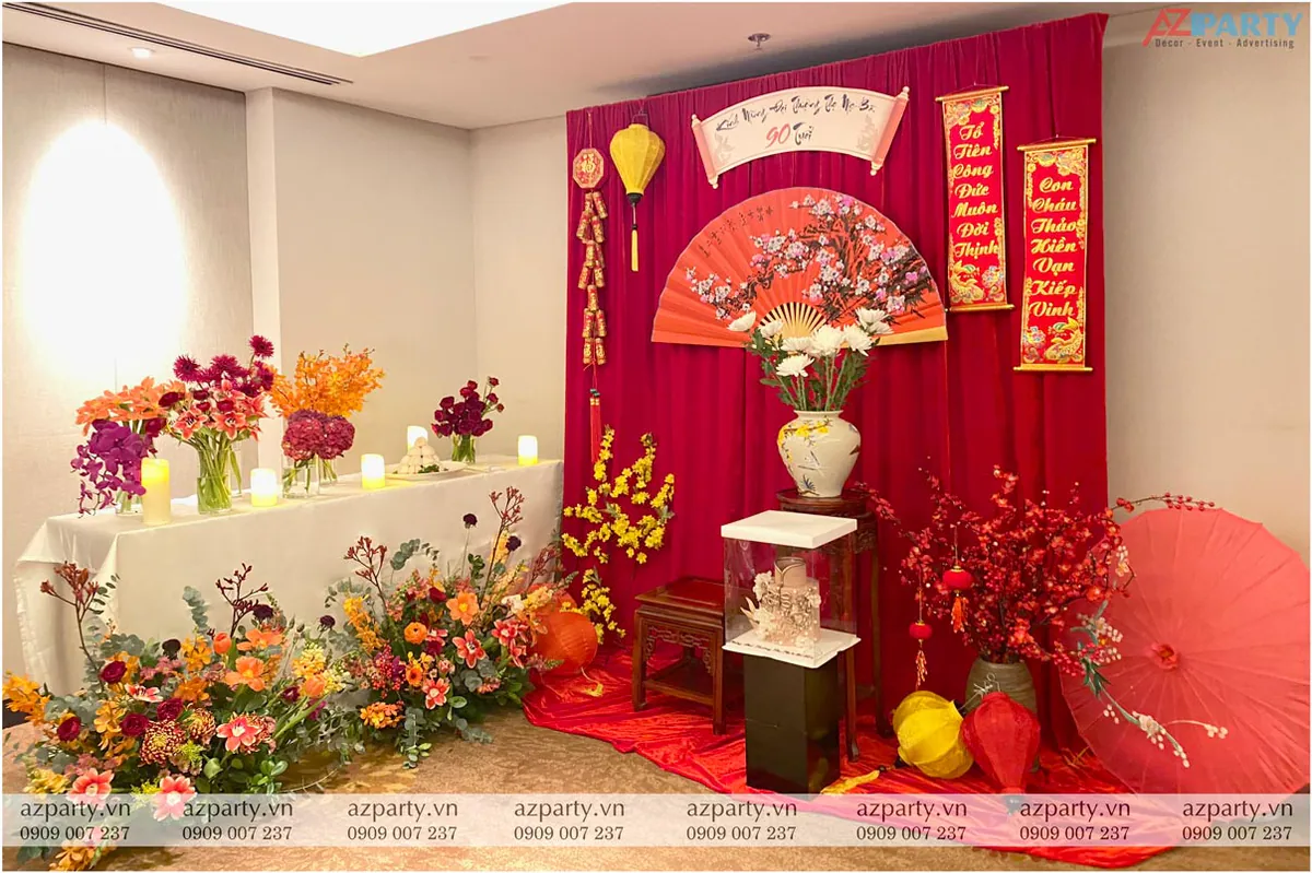 Khu vực sân khấu lễ thượng thọ cụ bà 90 tuổi với rèm nhung đỏ, đèn lồng vàng, quạt giấy vẽ cành đào và bàn gallery trưng bày hoa tươi rực rỡ.