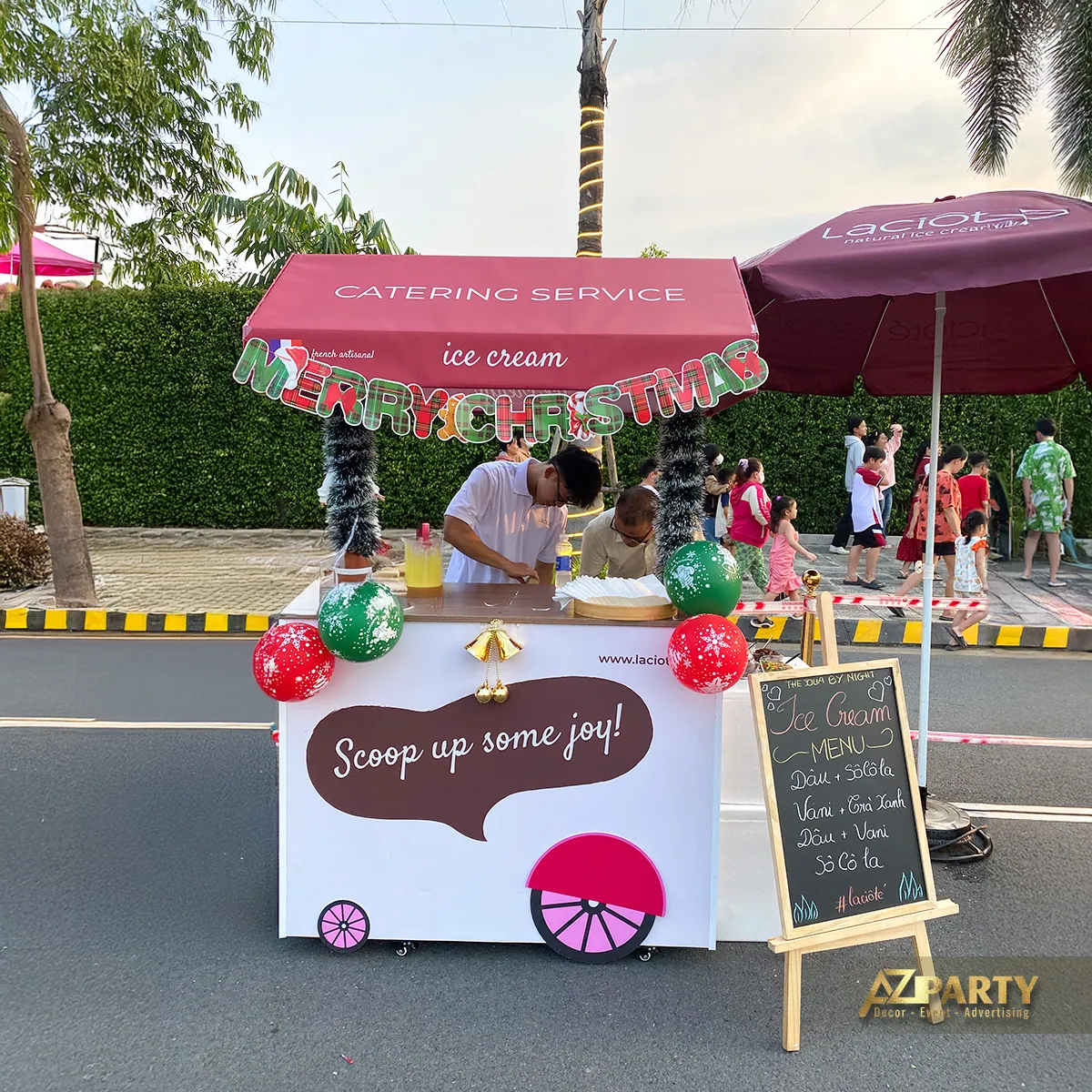 Quầy phục vụ nhỏ dưới ô màu hồng với bóng bay và khách tham gia sự kiện Noel
