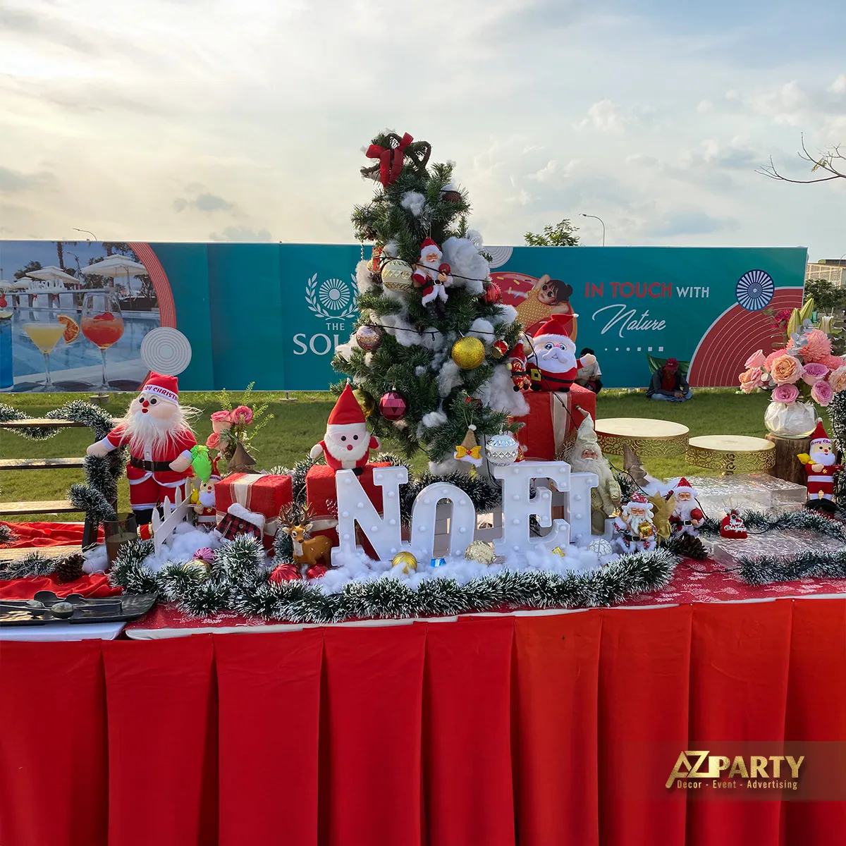 Tiểu cảnh Noel trên bàn phủ khăn đỏ với cây thông, ông già Noel và chữ NOEL lớn