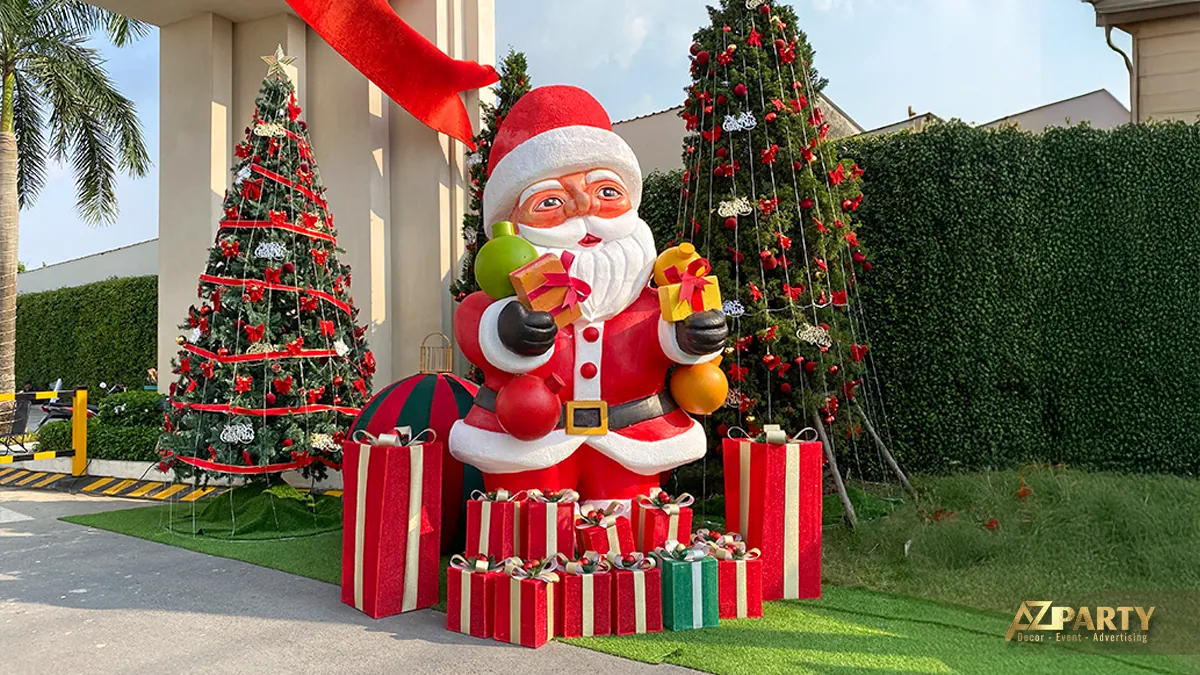Tượng ông già Noel cầm hộp quà, đứng giữa các hộp quà và cây thông trang trí