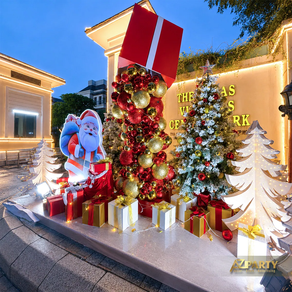 Tiểu cảnh Noel ban đêm tại khu vực cổng với cây thông, hộp quà, nhân vật xanh và dải nơ đỏ