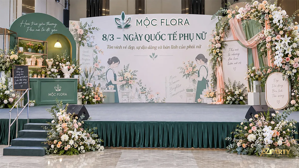 Phông nền xanh kem có hai người phụ nữ cắm hoa, quầy hoa bên trái và vòm hoa phối rèm bên phải