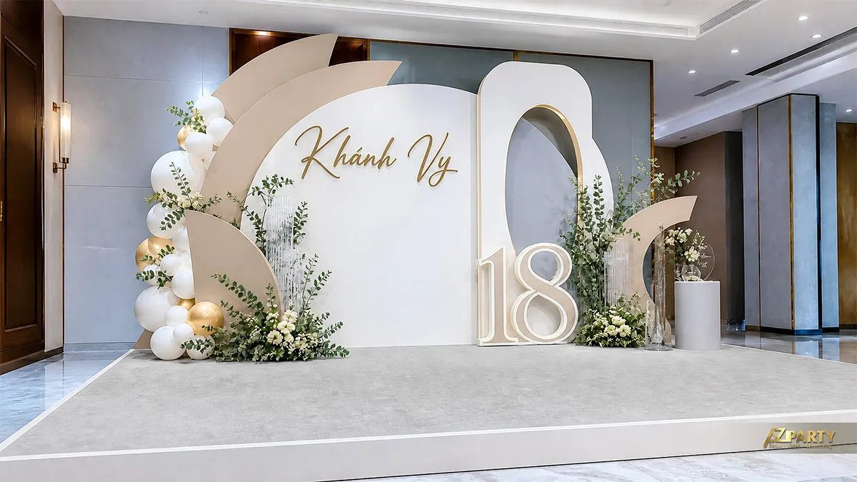Backdrop sinh nhật 18 tuổi cho Khánh Vy tông trắng kem gold, phối lá xanh, hoa và số 18.