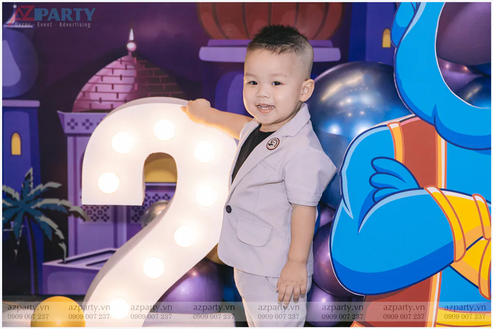 Bé Gia Hỷ diện bộ vest xám thanh lịch, cười rạng rỡ bên mô hình số 2 phát sáng tại sân khấu tiệc sinh nhật.