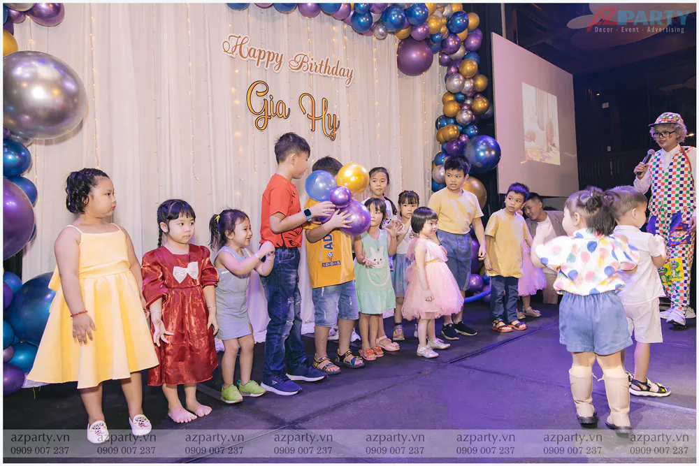 Chú hề trong trang phục nhiều màu sắc tổ chức các trò chơi hoạt náo vui tươi cho các bé trên sân khấu tiệc sinh nhật.