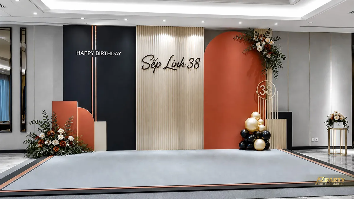 Backdrop sinh nhật sếp Linh 38 tuổi tông đen cam đất, phối vách lam, hoa và cụm bóng gold.