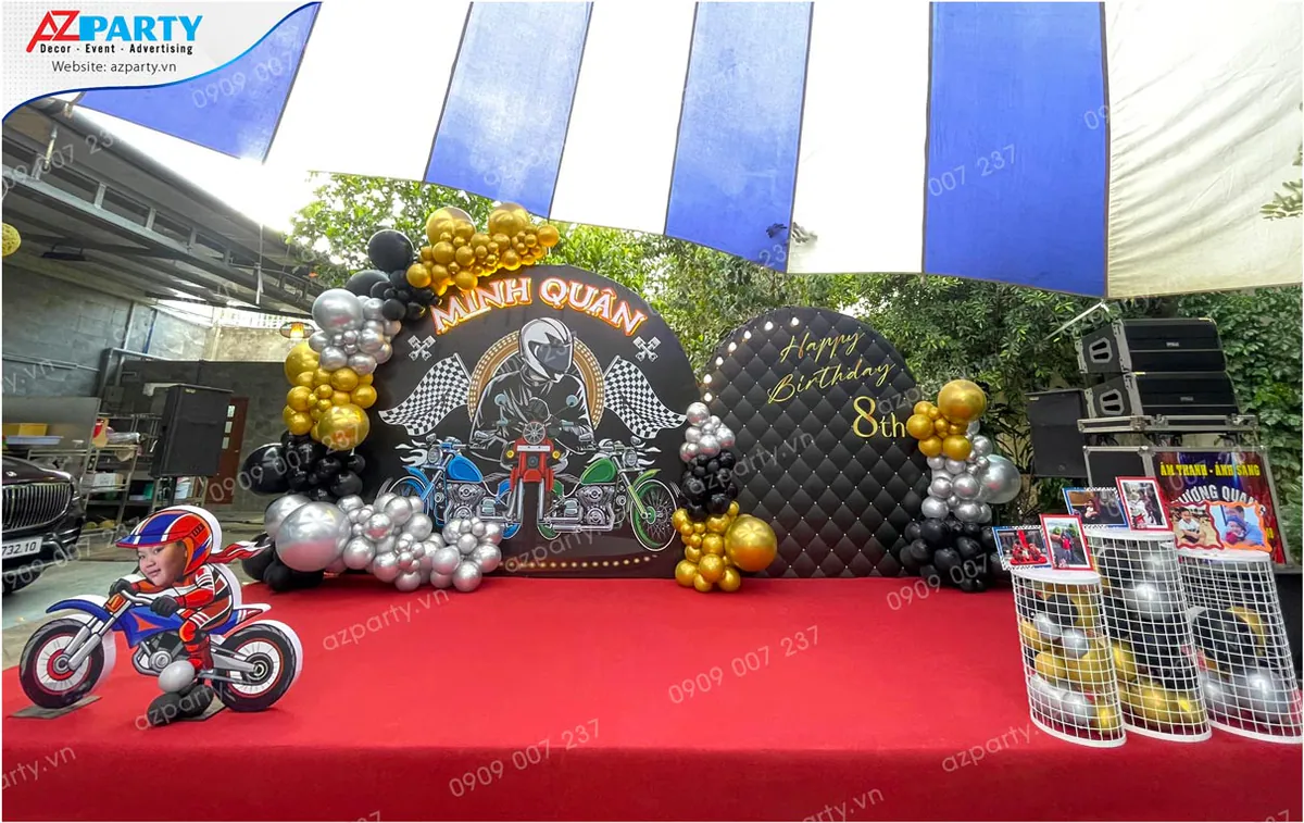 Trang trí tiệc sinh nhật ngoài trời chủ đề xe phân khối lớn cho bé Minh Quân tròn 8 tuổi với backdrop hình tay lái mô tô cực ngầu, thảm đỏ, mô hình chibi bé lái xe cùng bóng bay màu vàng, bạc và đen.