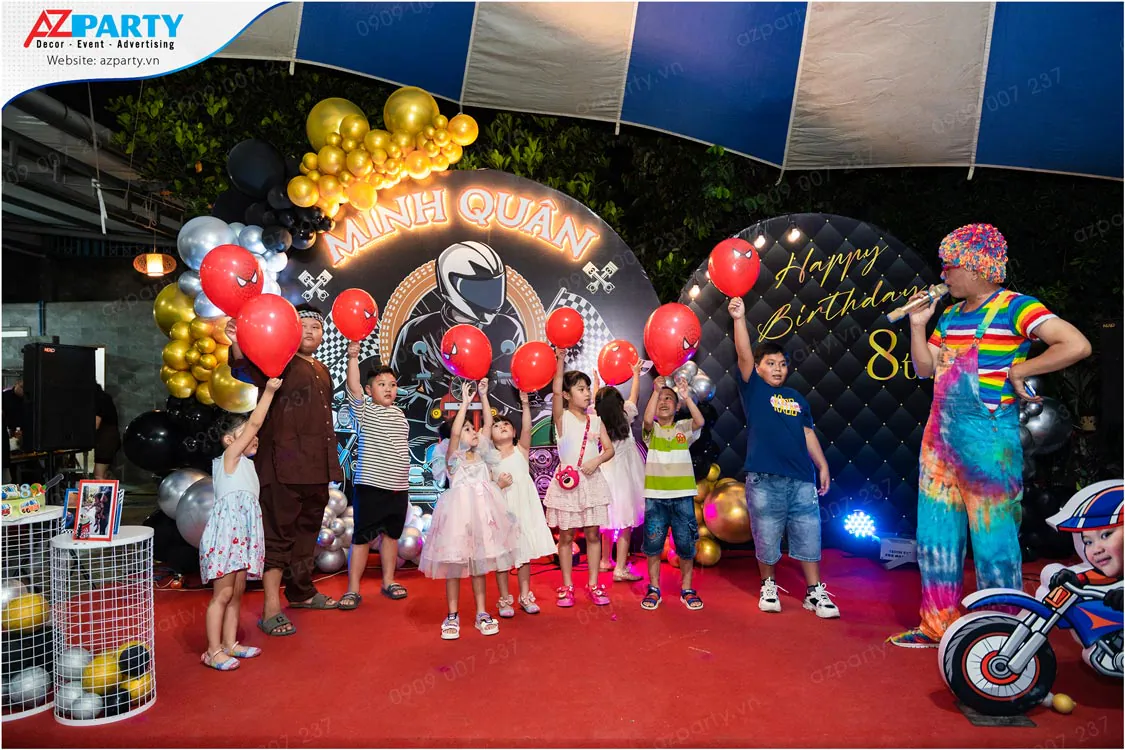 Các bé vui vẻ tham gia trò chơi sân khấu cùng chú hề MC bảy sắc cầu vồng, cầm trên tay những quả bóng bay màu đỏ in hình người nhện đáng yêu.