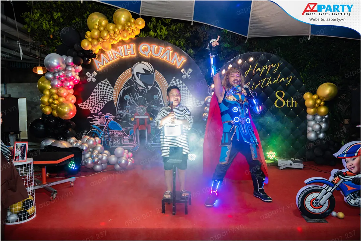 Nghệ sĩ hóa trang thành Thor (Thần Sấm) đang cùng bé Minh Quân biểu diễn màn xuất hiện đầy hoành tráng với búa Mjolnir phát sáng trên sân khấu tiệc sinh nhật.
