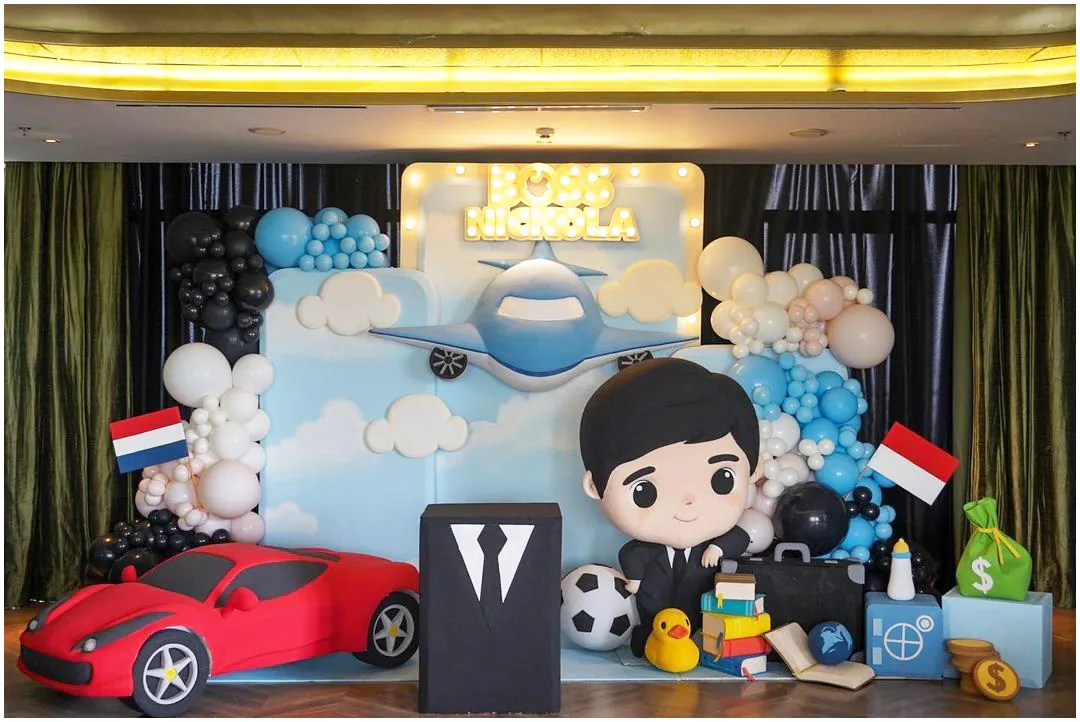 Trang trí tiệc thôi nôi bé Nickola với chủ đề Boss Baby đầy độc đáo, có mô hình máy bay màu xanh, xe ô tô màu đỏ, hình chibi bé mặc vest lịch lãm và bóng bay tone màu xanh dương, trắng, đen.