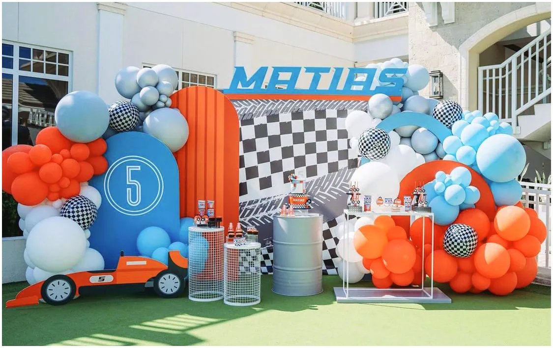 Trang trí tiệc sinh nhật ngoài trời cho bé Matias tròn 5 tuổi với chủ đề xe đua thể thao năng động, sử dụng cờ đua carô trắng đen làm nền, mô hình xe công thức 1 màu cam và chùm bóng bay phối màu cam, xanh dương, trắng.