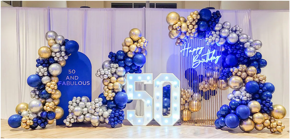 Trang trí tiệc sinh nhật 50 tuổi (50 and Fabulous) hoành tráng với chữ số 50 gắn đèn LED lớn ở trung tâm, kết hợp cổng bóng bay nghệ thuật tông màu xanh dương, vàng và bạc sang trọng dưới ánh đèn neon xanh 'Happy Birthday'.