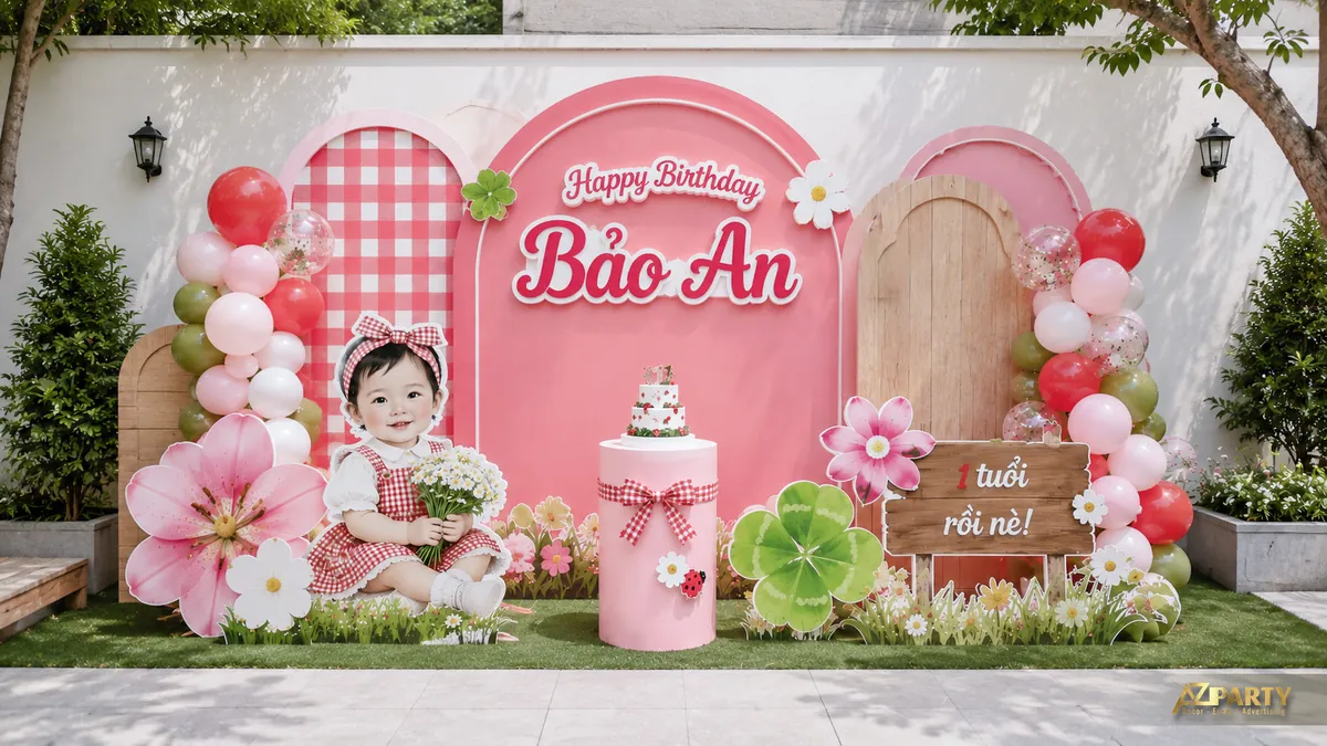 Backdrop caro hồng cho bé Bảo An với hoa lớn, cỏ xanh và bàn bánh đặt giữa