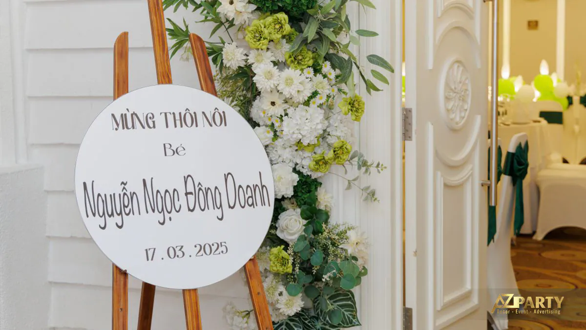 Bảng chào mừng thôi nôi bé Nguyễn Ngọc Đông Doanh đặt trên giá đỡ gỗ trang trí hoa tươi