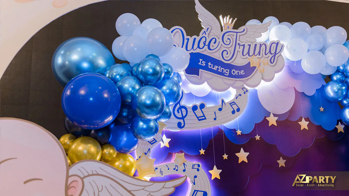 Bảng chữ Quốc Trung Is turning one được cách điệu mềm mại bên cạnh dải bóng bay xanh dương và các nốt nhạc vui tươi
