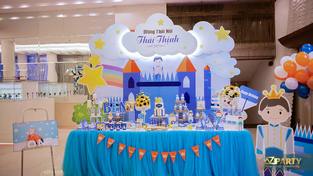 Góc chính diện bàn tiệc thôi nôi bé Thái Thịnh nổi bật với hình ảnh chibi hoàng tử nhỏ và bảng ảnh thôi nôi của bé