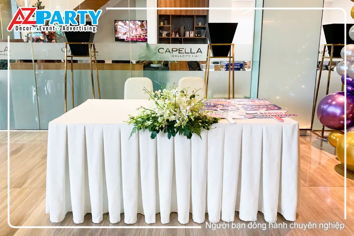 Bàn đón khách trải khăn trắng trang trí hoa tươi tươi tắn và các bảng hashtag check-in sự kiện Year End Party của Shinryo tại Capella Gallery Hall.
