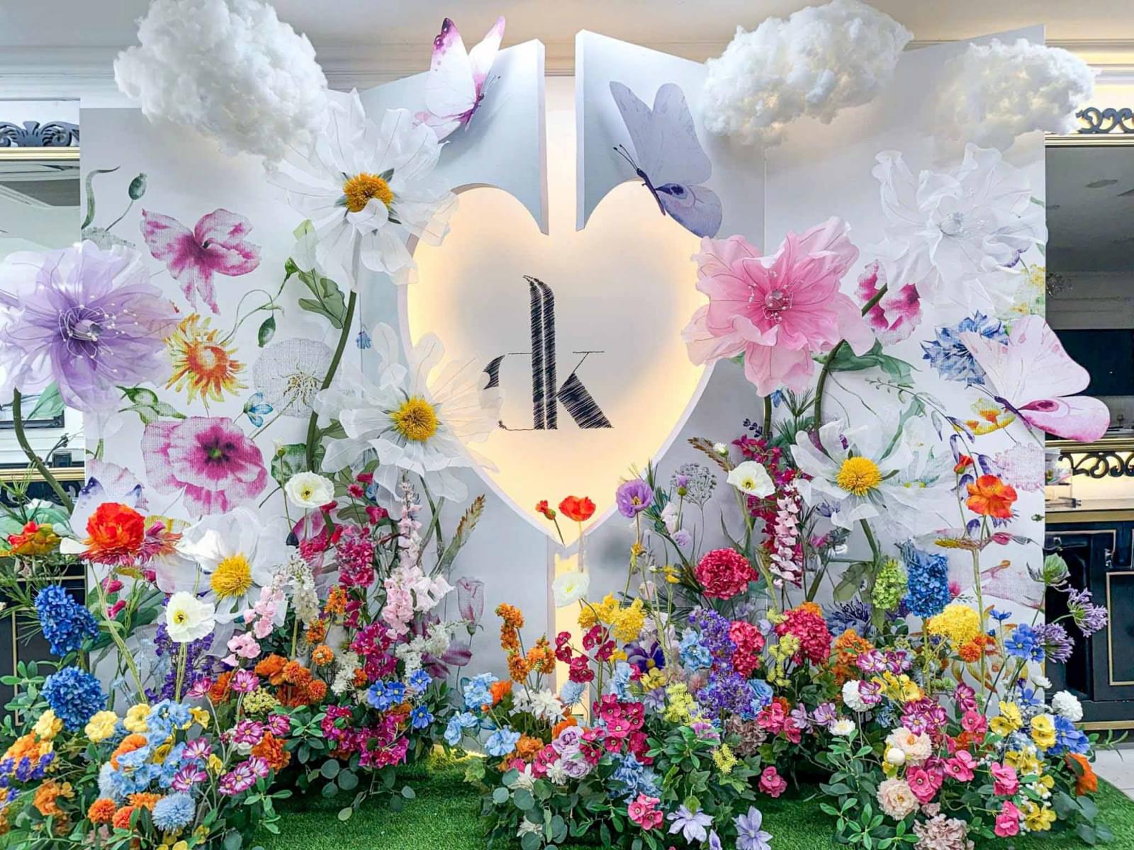 Backdrop trang trí 8/3 phong cách khu vườn cổ tích với khung trái tim phát sáng ở trung tâm, nền trắng phối họa tiết hoa và bướm bay nhẹ nhàng.