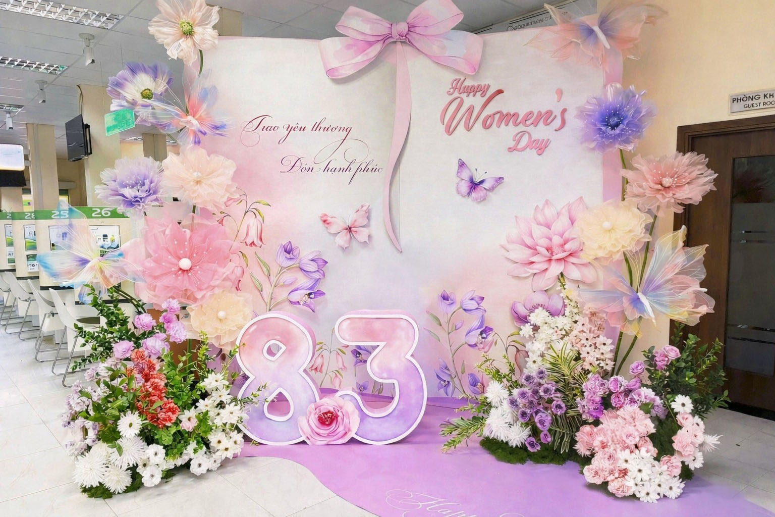 Backdrop 8/3 tone hồng pastel với nơ lớn, bộ số 8.3 và hoa van trang trí, bươm bướm lấp lánh tại văn phòng công ty