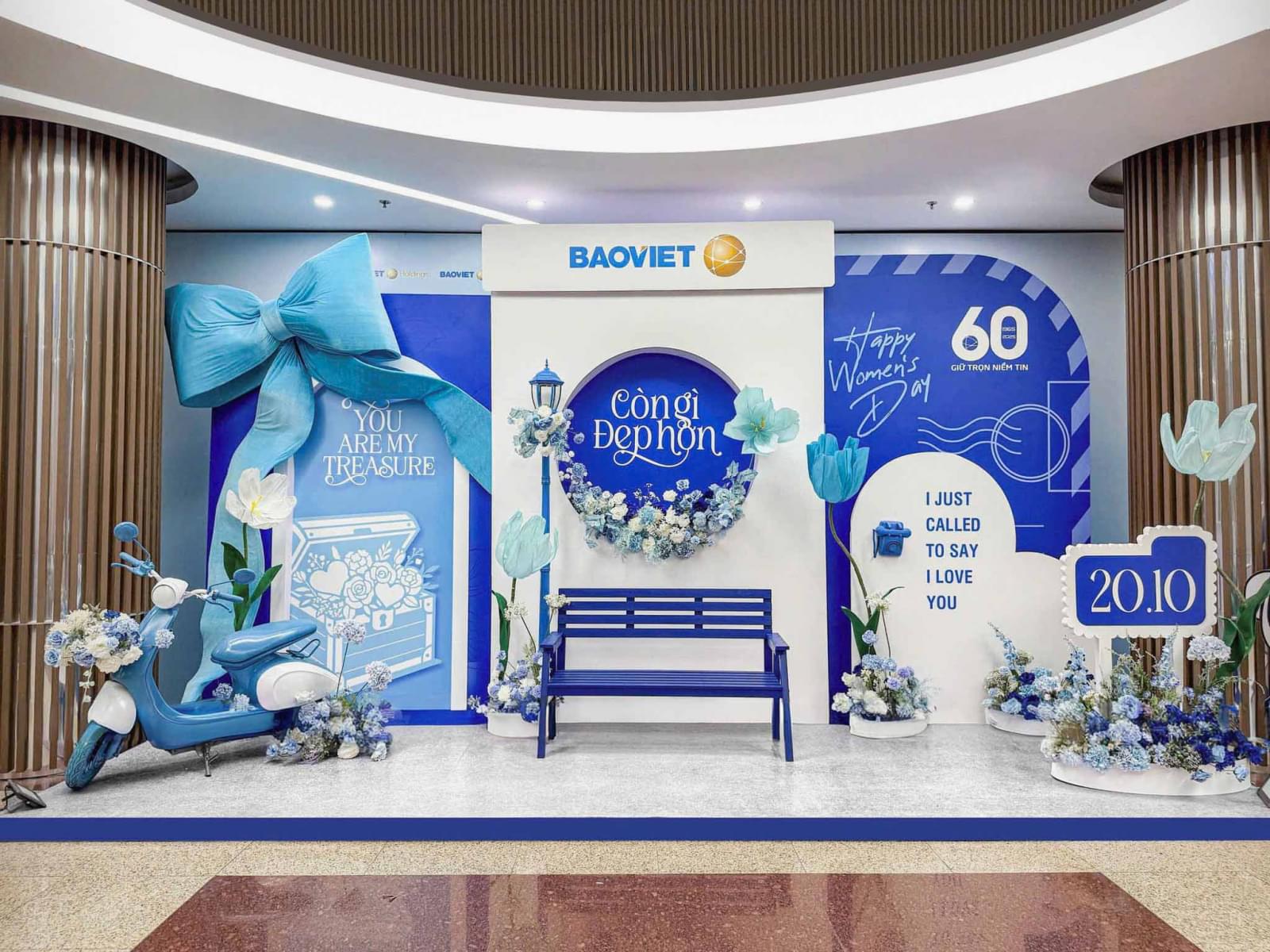 Backdrop 20/10 tone xanh dương sang trọng với chi tiết nơ lớn, ghế băng, xe scooter và hoa trang trí trong không gian văn phòng.