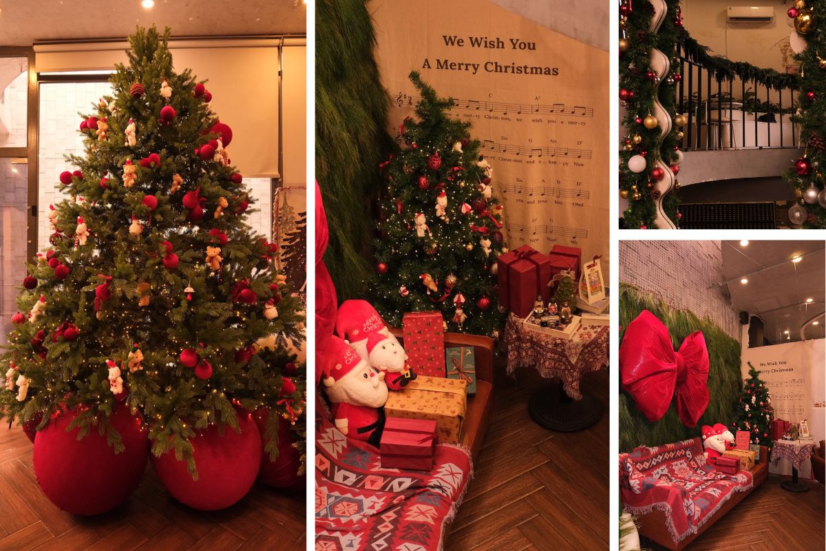 Góc trang trí Giáng sinh ấm cúng với cây thông xanh treo đầy quả châu đỏ, ghế sofa phủ khăn họa tiết và nơ lớn trang trí trên tường. Không gian được tô điểm bằng hộp quà, ông già Noel bông và phông nền in lời bài hát “We Wish You A Merry Christmas”, tạo cảm giác gần gũi và lễ hội.