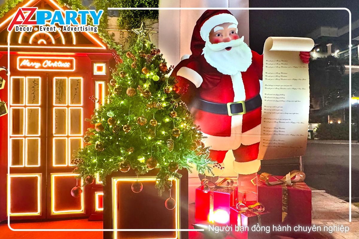 cận cảnh cây thông noel với quả châu đỏ cùng dây đèn vàng, bên cạnh là ông già noel vác quà, tay còn lại cầm danh sách các trẻ em được nhận quà tặng