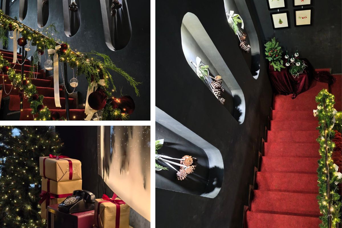 Cầu thang với thảm đỏ và tường đen được trang trí Giáng sinh bằng dây đèn, quả thông, nơ nhung và hộp quà lớn. Ánh sáng vàng ấm kết hợp chi tiết trang trí tối giản tạo nên không gian sang trọng, cổ điển và ấn tượng