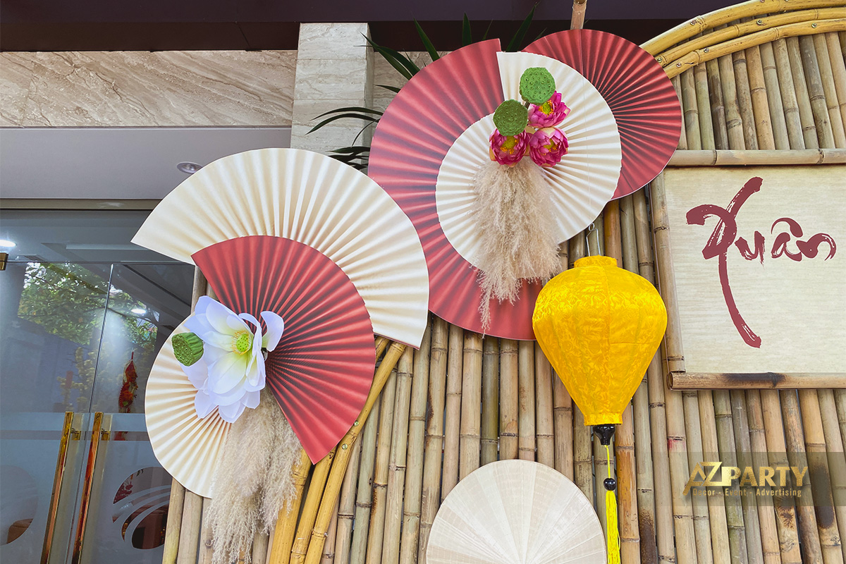 chi tiết trang trí Tết với quạt giấy đỏ kem, hoa sen trắng, hoa mẫu đơn hồng và đèn lồng vàng treo trên nền tre trúc 