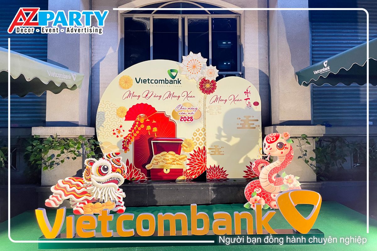 Tiểu cảnh tết tại Ngân hàng Vietcombank - Chi nhánh Sài Thành, phía trước là hộp logo 3D logo của ngân hàng, phía sau là mô hình chú lân , phía còn lại là mô hình rắn linh vật 2025, sau lưng là backdrop với tone màu vàng sữa 